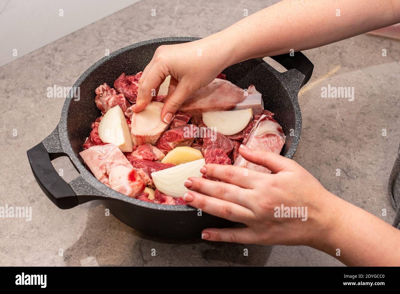 Frau Hände Bereiten Rindfleisch Auf Küche. Nahaufnahme des Kochens des traditionellen jüdischen Cholent Hamin Hauptgericht für den Schabbat. Marrowbone wird einem Topf mit Kartoffeln, Zwiebeln, Bohnen und Weizenkörnern hinzugefügt. Stockfoto
