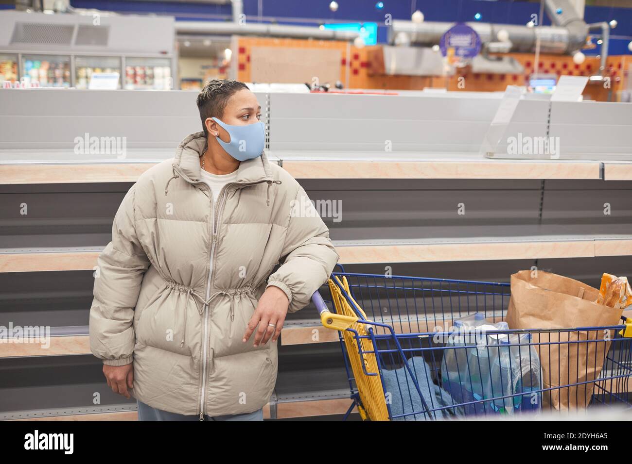 Junge Frau in Schutzmaske stehend mit Warenkorb in Supermarkt und Einkaufen Stockfoto