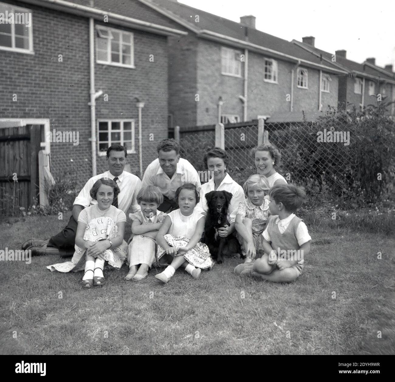 1950er Jahre, historisch, in einem Garten hinter einigen Doppelhaushälften Backstein-Häuser, sitzen zwei Familien zusammen für ein Gruppenfoto England, Großbritannien. Großbritannien nach dem Krieg war ein unschuldigeres, weniger kompliziertes Alter, in dem elegant aussehende Frauen glücklich waren, Mütter zu sein und Kinder aussahen, also Kinder. Stockfoto