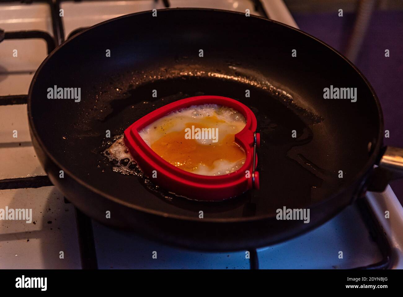 Pfannengebratenes Ei in Form eines Herzens auf einem weißen Gasherd und schwarzem Grill, der die Pfanne über einer Feuerflamme hält Stockfoto