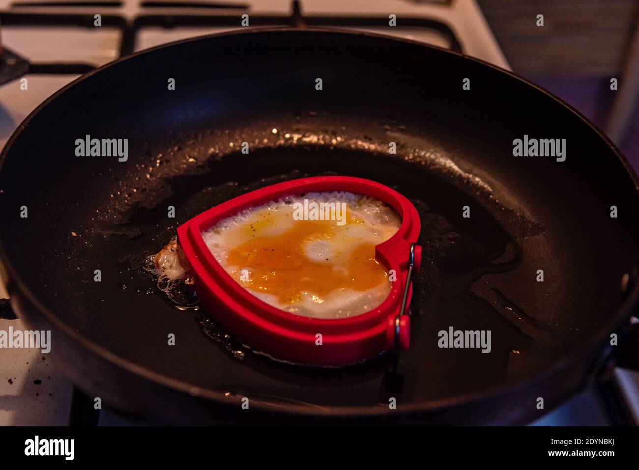 Pfannengebratenes Ei in Form eines Herzens auf einem weißen Gasherd und schwarzem Grill, der die Pfanne über einer Feuerflamme hält Stockfoto