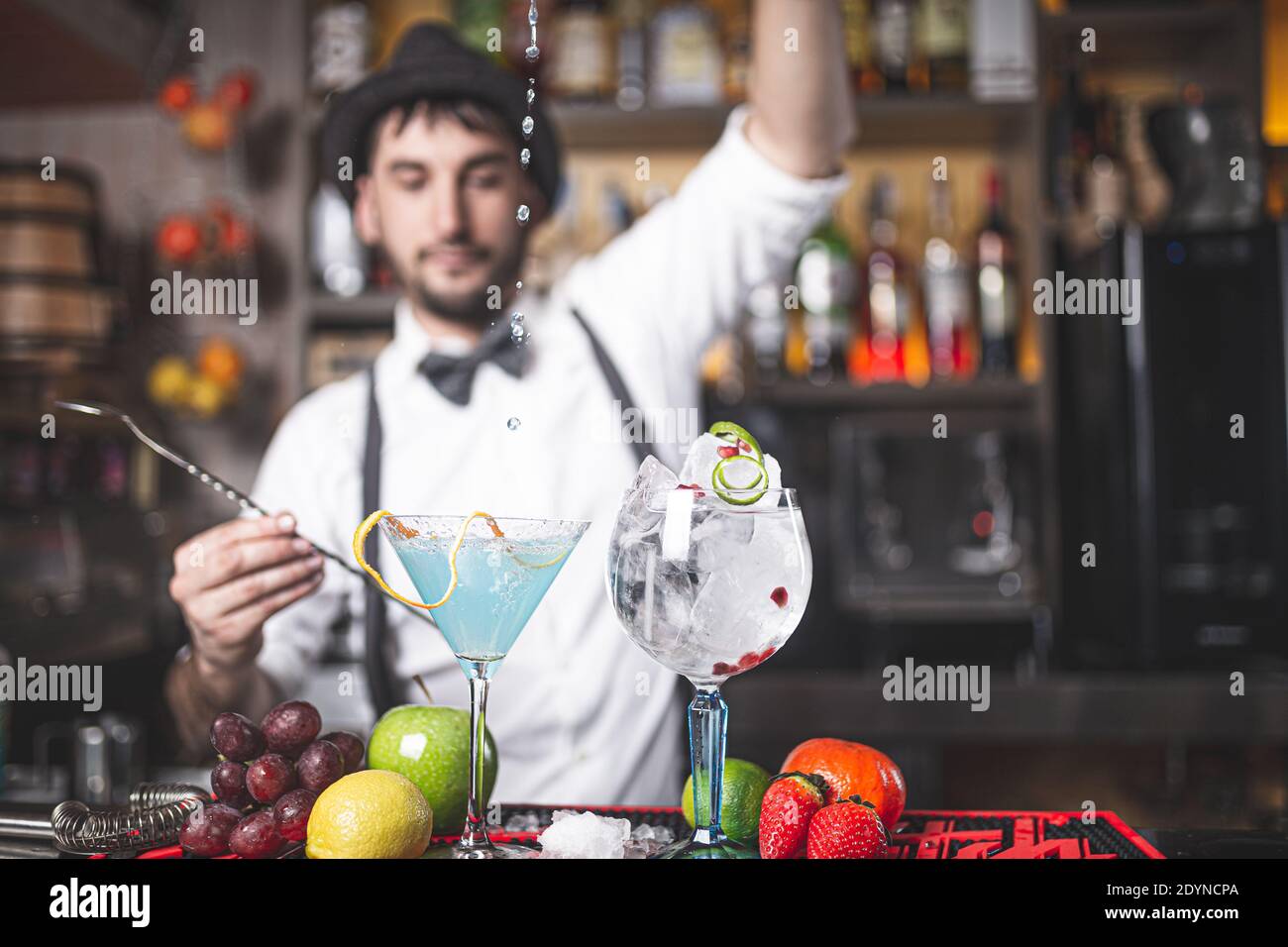 Der erfahrene Barkeeper macht im Nachtclub einen Cocktail mit Hut und Fliege. Tropfen fallen in den Becher. Stockfoto