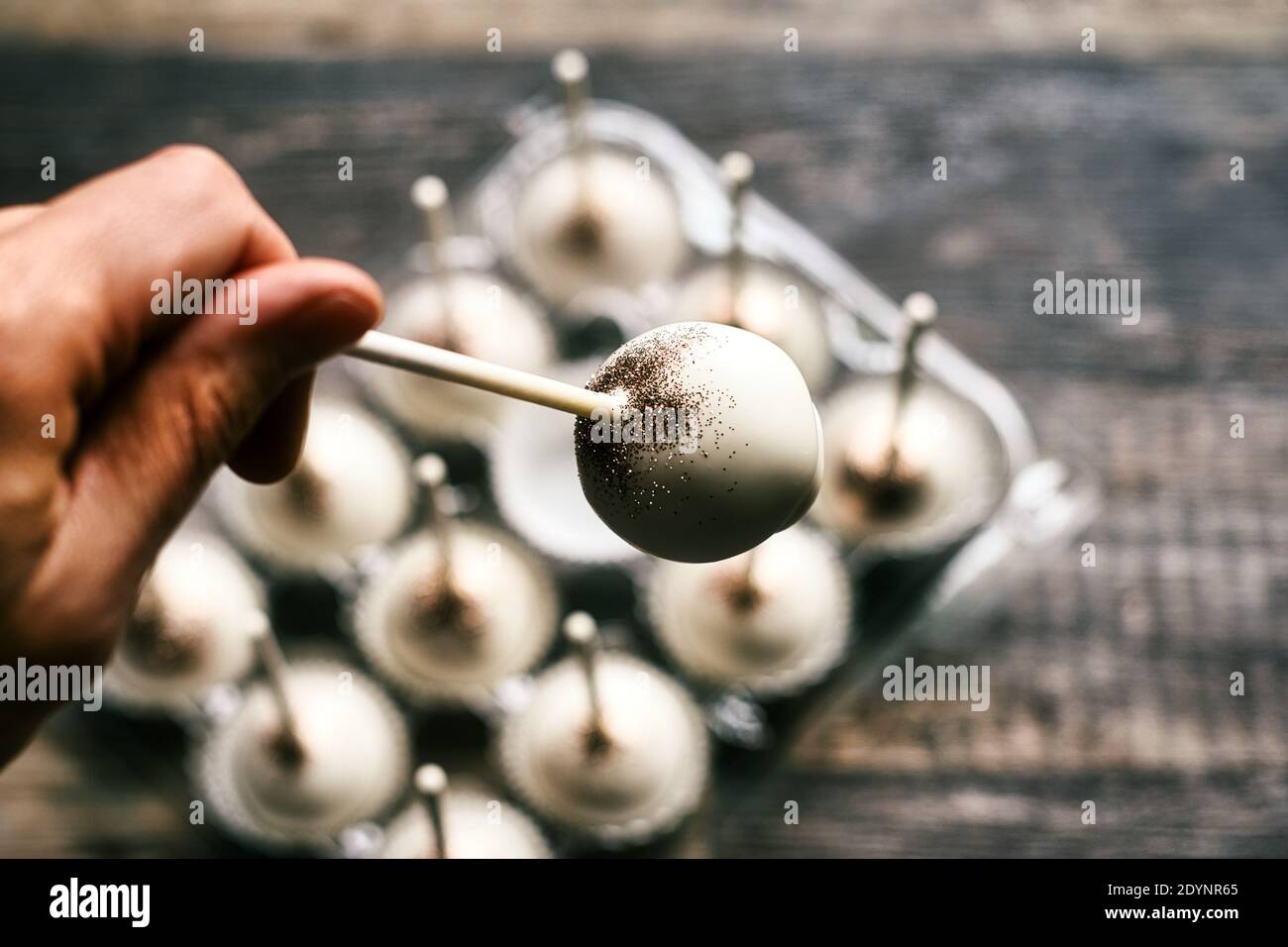 Die Hand eines Bäckers hält einen Cake Pop aus einer Reihe von glitzernden Kuchenknallen auf einem Holztisch. Stockfoto
