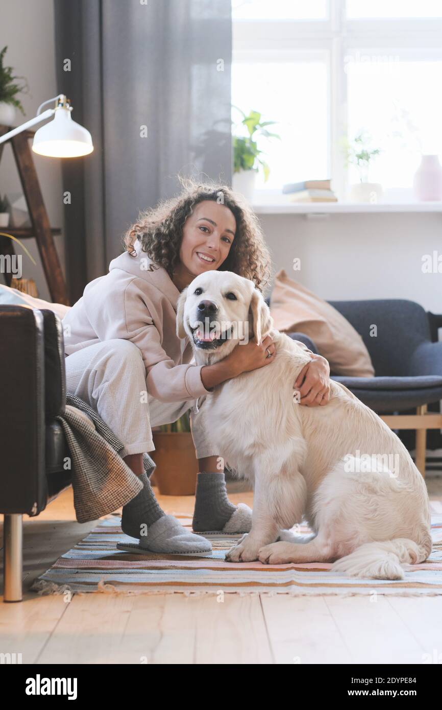 Porträt einer glücklichen jungen Frau, die während des Sitzens die Kamera anlächelt Auf dem Boden im Zimmer und umarmt ihren Hund Stockfoto
