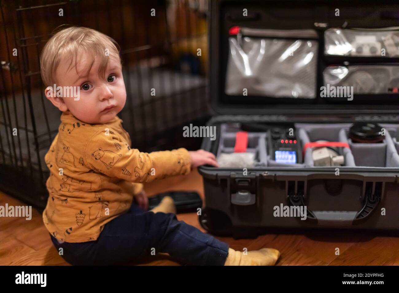 Netter kleiner Junge, der die Kamera anschaut, während er den Koffer seines Vaters berührt, der mit professioneller Videoausrüstung gefüllt ist. Neugierde und Entdeckungskonzept. Stockfoto