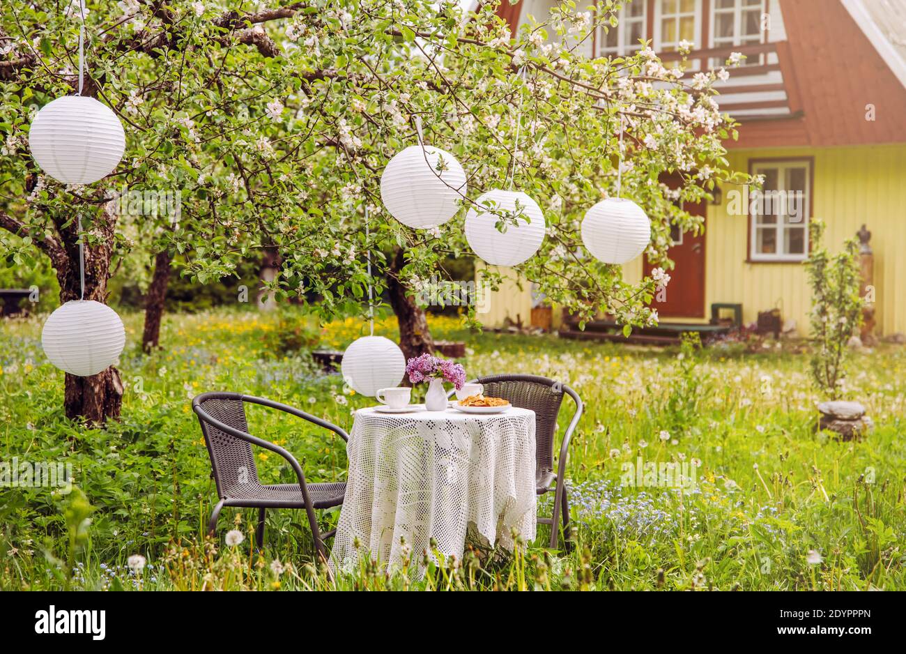 Terrassenmöbel im Apfelgarten auf wildem, langen Rasen, Tisch mit weißer Tischdecke und weißen Papierlaternen, die vom Apfelbaum hängen. Niedliches Holz. Stockfoto
