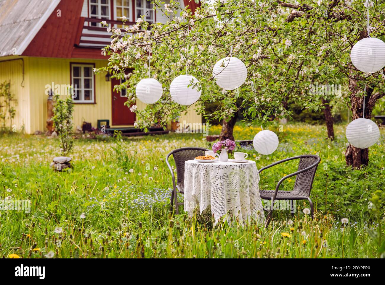 Terrassenmöbel im Apfelgarten auf wildem, langen Rasen, Tisch mit weißer Tischdecke und weißen Papierlaternen, die vom Apfelbaum hängen. Niedliches Holz. Stockfoto