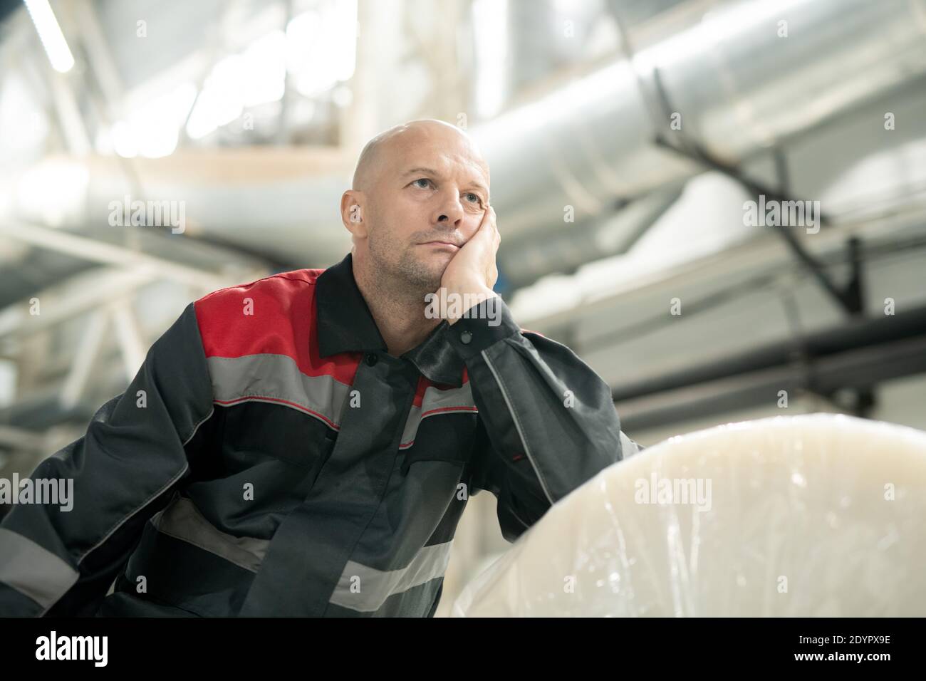 Junge nachdenkliche Glatze Vorarbeiter in Arbeitskleidung lehnt auf riesige Paket Von gerollten Polyethylen-Folie beim Nachdenken über Arbeitsfragen Stockfoto