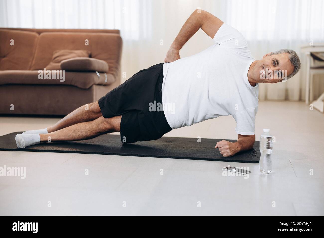 Fröhlicher älterer Mann, der auf einer Yogamatte eine seitliche Plank-Übung macht. Wie man gesund auf Quarantäne-Konzept zu bleiben. Gesunder Lebensstil. Athletic Mann Training und Stockfoto