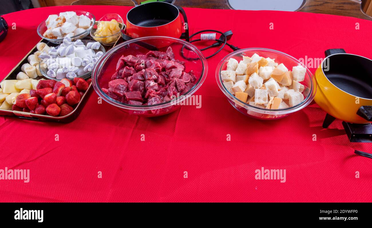 Verschiedene Speisen für Fondue, darunter Fleisch, französisches Brot geschnitten und Dessert mit Erdbeeren, Äpfeln, Banane, Angel Food Kuchen Ananas und Marshmallows Stockfoto