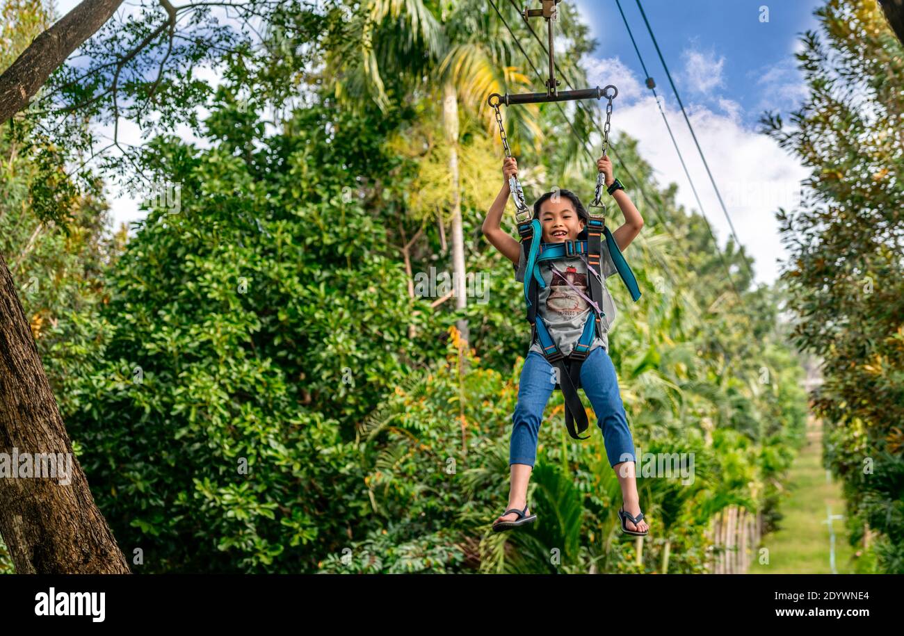 Asiatische tapfere Kind Mädchen ziplining zwischen großen Bäumen, lächelndes Gesicht, Tageszeit extreme Outdoor-Aktivität in Thailand. Mit medizinischen Maske mit Gurt hängt Stockfoto