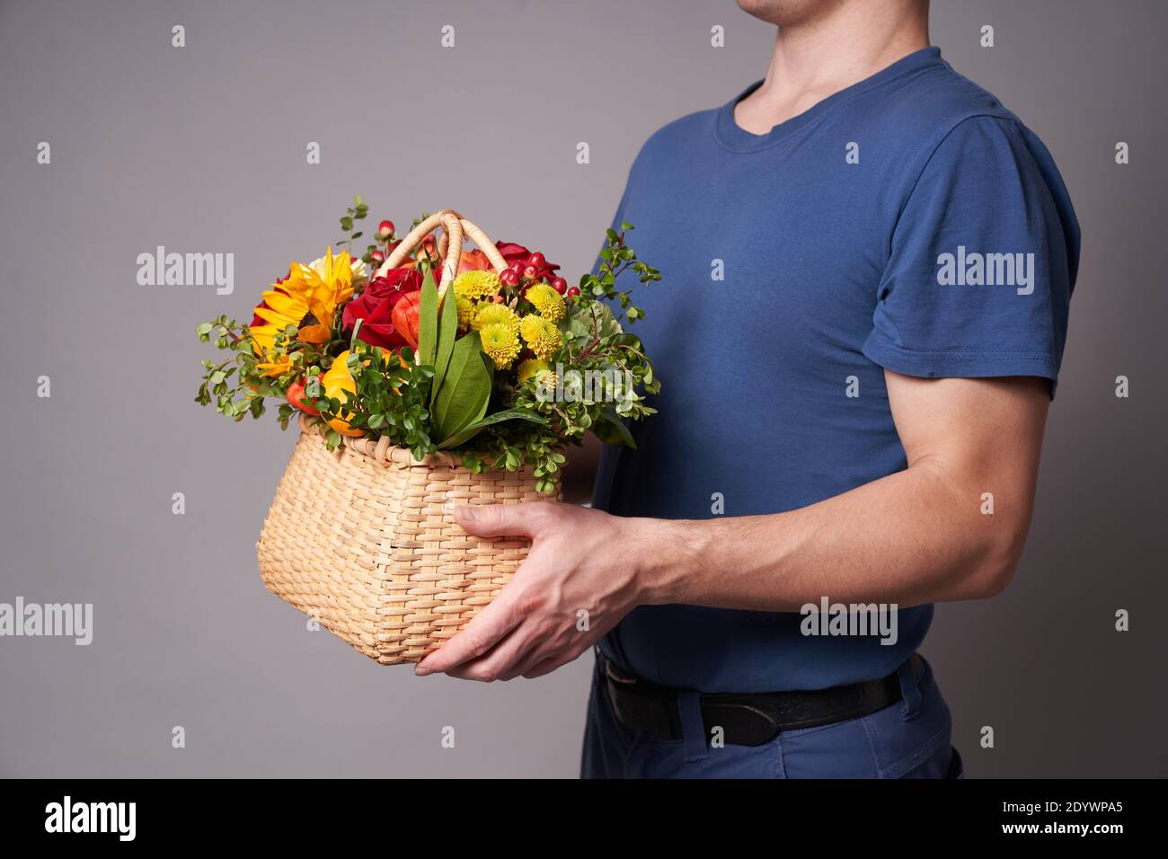 Ein weißer Mann in einem blauen T-Shirt hält einen Blumenkorb mit einem Kopierplatz auf einem grauen Hintergrund, Blumenlieferung oder ein Geschenk für den Urlaub Stockfoto