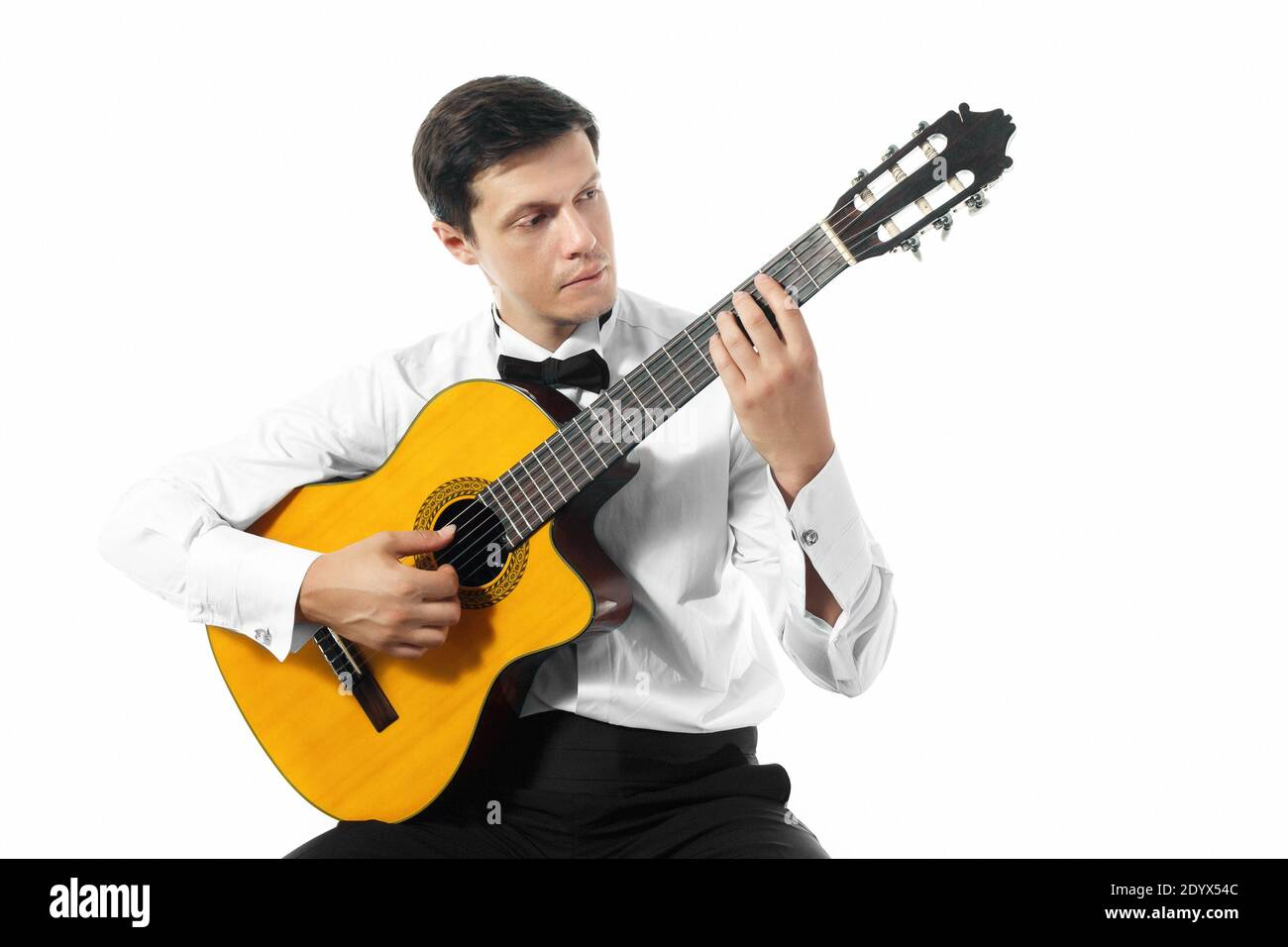 Junger Mann in weißem Hemd mit Fliege posiert mit klassischer Gitarre im Studio auf weißem Hintergrund. Stockfoto