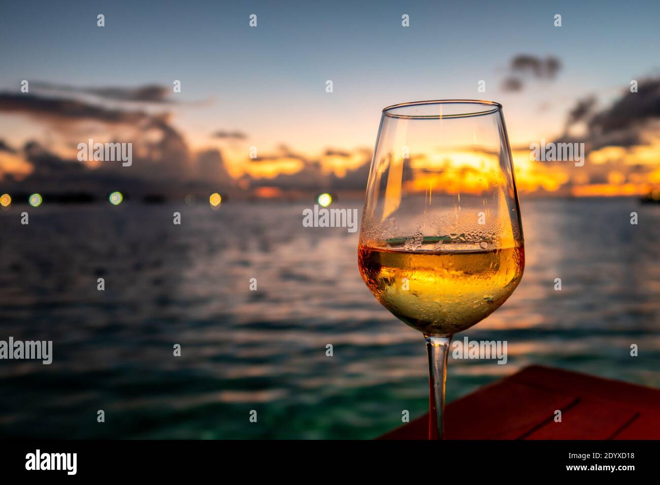 Glas Weißwein auf einem Tisch mit dramatischen Sonnenuntergang im Hintergrund und Lichter am Horizont, Malediven. Stockfoto
