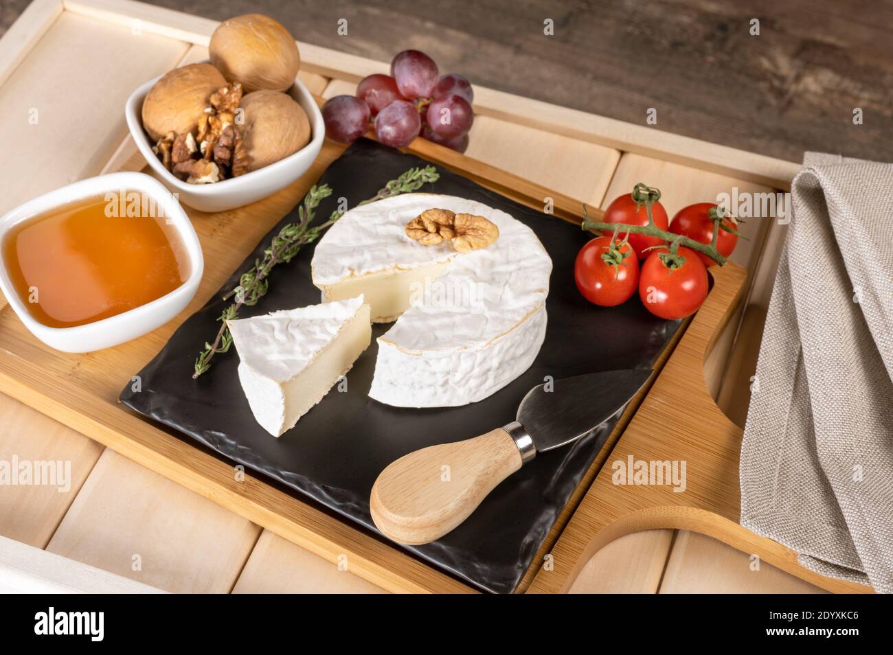 Holztablett zum Frühstück mit Brie-Käse, Honig, Walnüssen, Tomaten. Käse Frühstück auf dem Holztablett mit schwarzem Teller Stockfoto