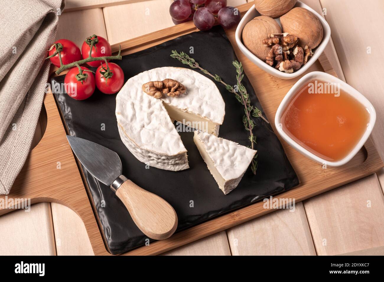 Holztablett zum Frühstück mit Brie-Käse, Honig, Walnüssen, Tomaten. Käse Frühstück auf dem Holztablett mit schwarzem Teller Stockfoto