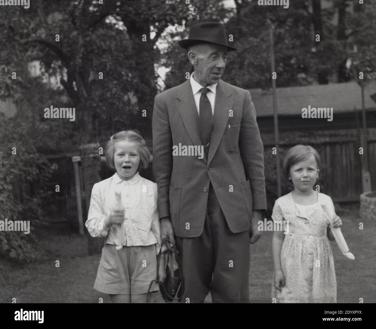 1950er Jahre, historisch, zwei junge Mädchen mit ihrem Großvater, der Anzug und Krawatte und einen dreifachen Hut trägt. Die jungen Mädchen tragen etwas, das aussieht wie Steinstöcke, ein beliebtes Süßes in dieser Zeit. Stockfoto