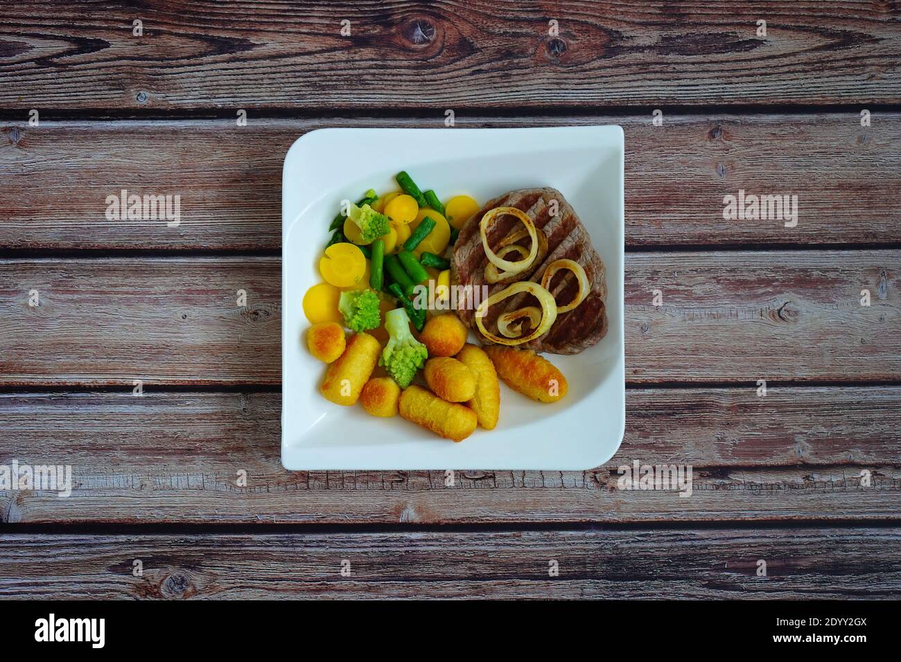 Rindersteak mit Kartoffelkroketten und Gemüse (grüner Spargel und gelbe Karotten) auf einem weißen quadratischen Teller mit dunklem Holzhintergrund. Stockfoto
