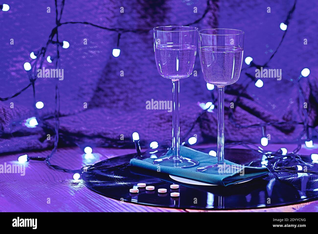 Selektives Fokusfoto von zwei Gläsern Champagner und Pillen auf dem Hintergrund einer Schallplatte, umgeben von heller Clubbeleuchtung Stockfoto