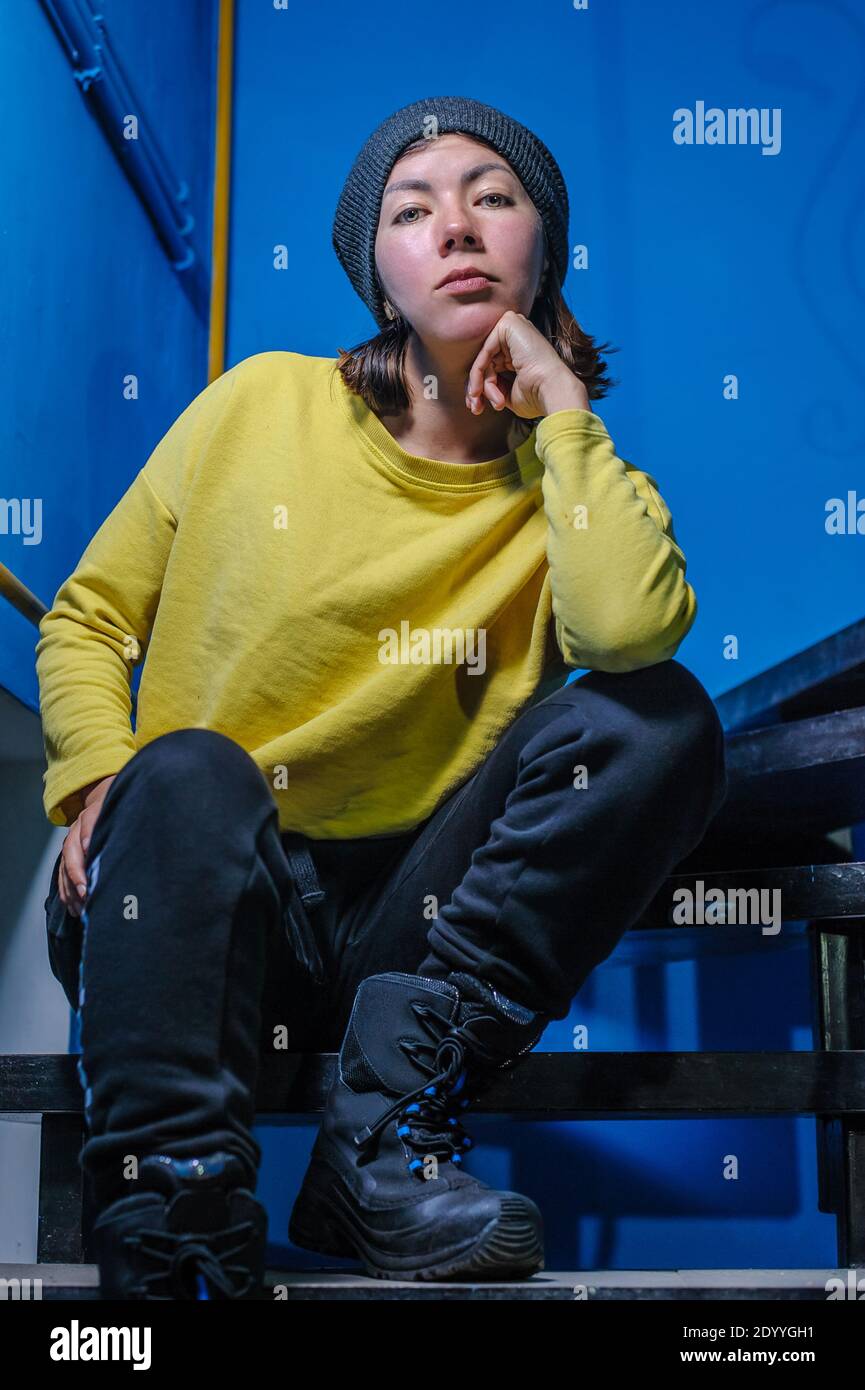 Teenager-Mädchen in einem warmen Winter Pullover Hoodie, Strickmütze und High-Sports-Schuhe sitzt auf den Stufen der Treppe auf einem blauen Hintergrund Stockfoto