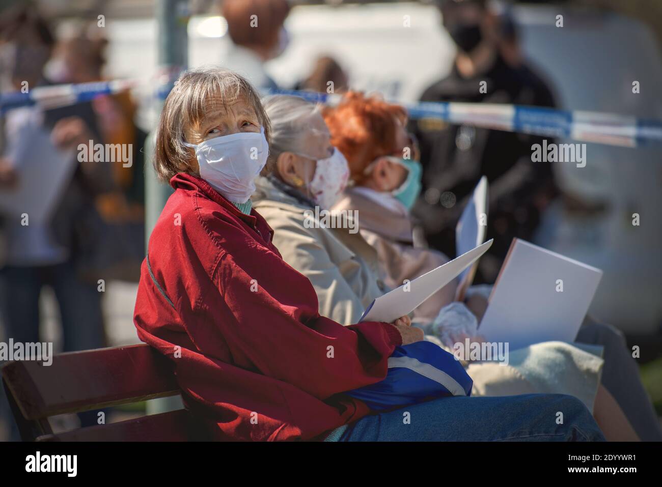 Drei ältere Frauen mit Gesichtsmasken sitzen auf einer Bank neben einer Schlange von Menschen warten auf einen Corona-Virus-Test. Stockfoto