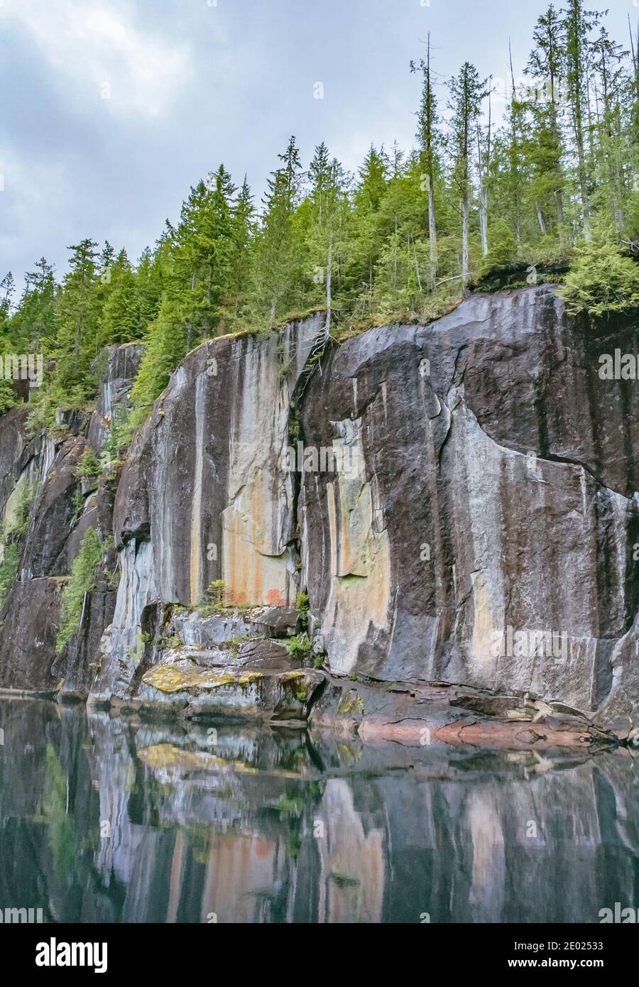 Gekrönt von dichtem Wald spiegeln sich steile senkrechte Klippen im stille Wasser darunter. Ein verblassenes Piktogramm ist sichtbar (Alison Sound, British Columbia). Stockfoto