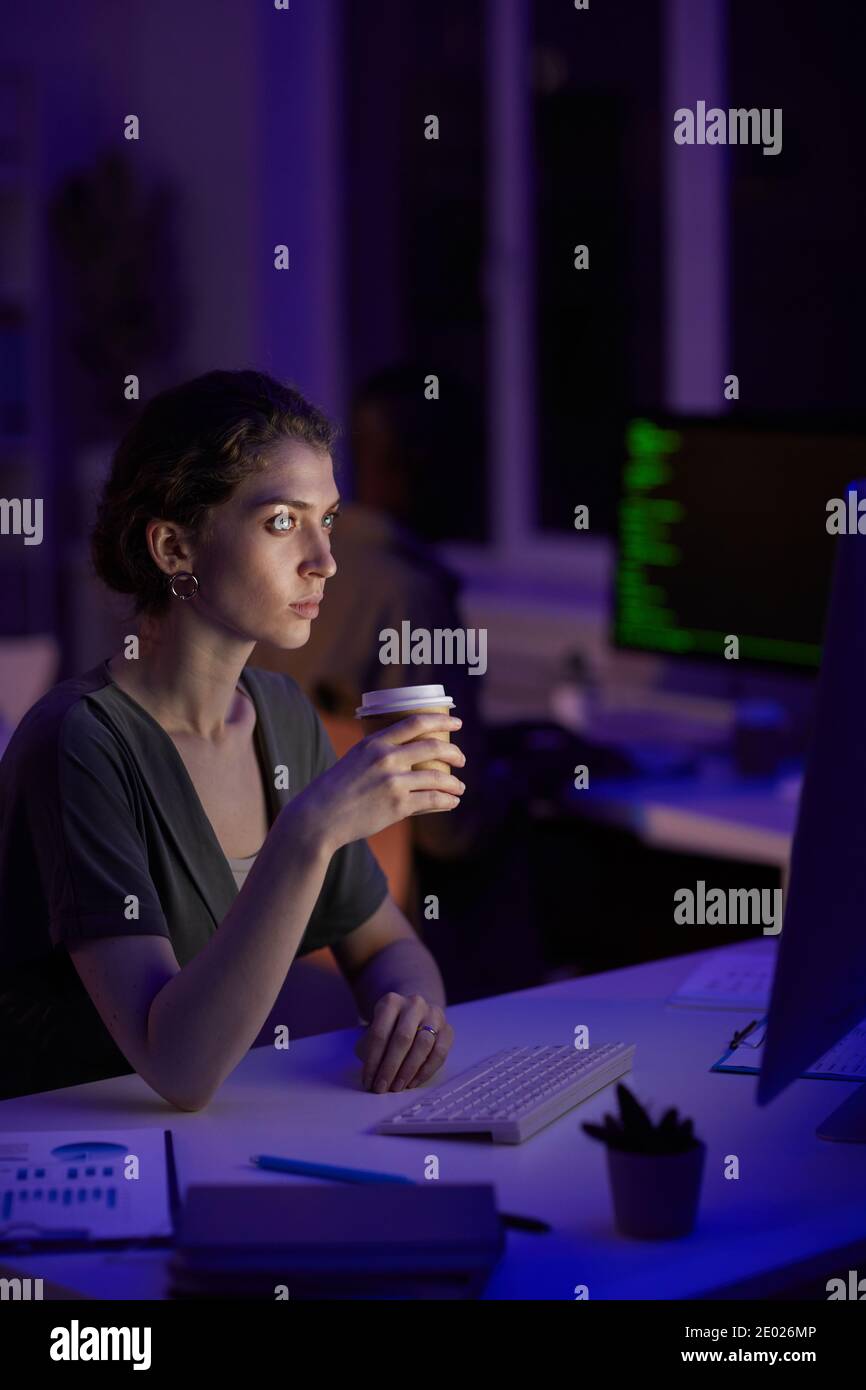 Moderne junge Frau sitzt in dunklen Bürozimmer spät in Abends Kaffee trinken Blick auf Computer-Monitor Stockfoto