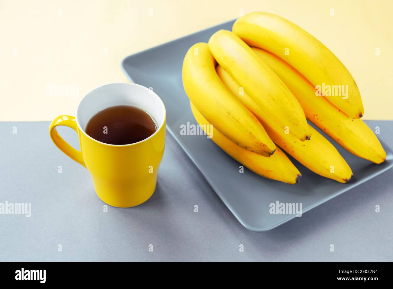 Gelbe Tasse Tee und Bananen auf grauem Teller. Trendige Farben 2021 Stockfoto