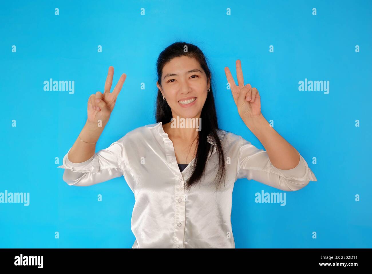 Eine attraktive Thailänderin, die sich glücklich fühlt, lächelt und die Hände hochhält und mit den Fingern das Siegeszeichen streicht. Einfacher hellblauer Hintergrund. Stockfoto