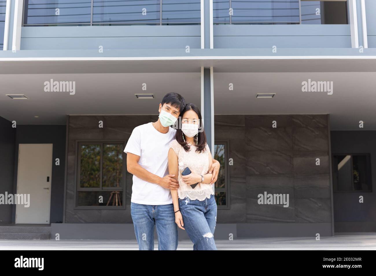 Asiatisches Paar mit Schutzmaske stehen und umarmen vor dem neuen Haus. Stockfoto