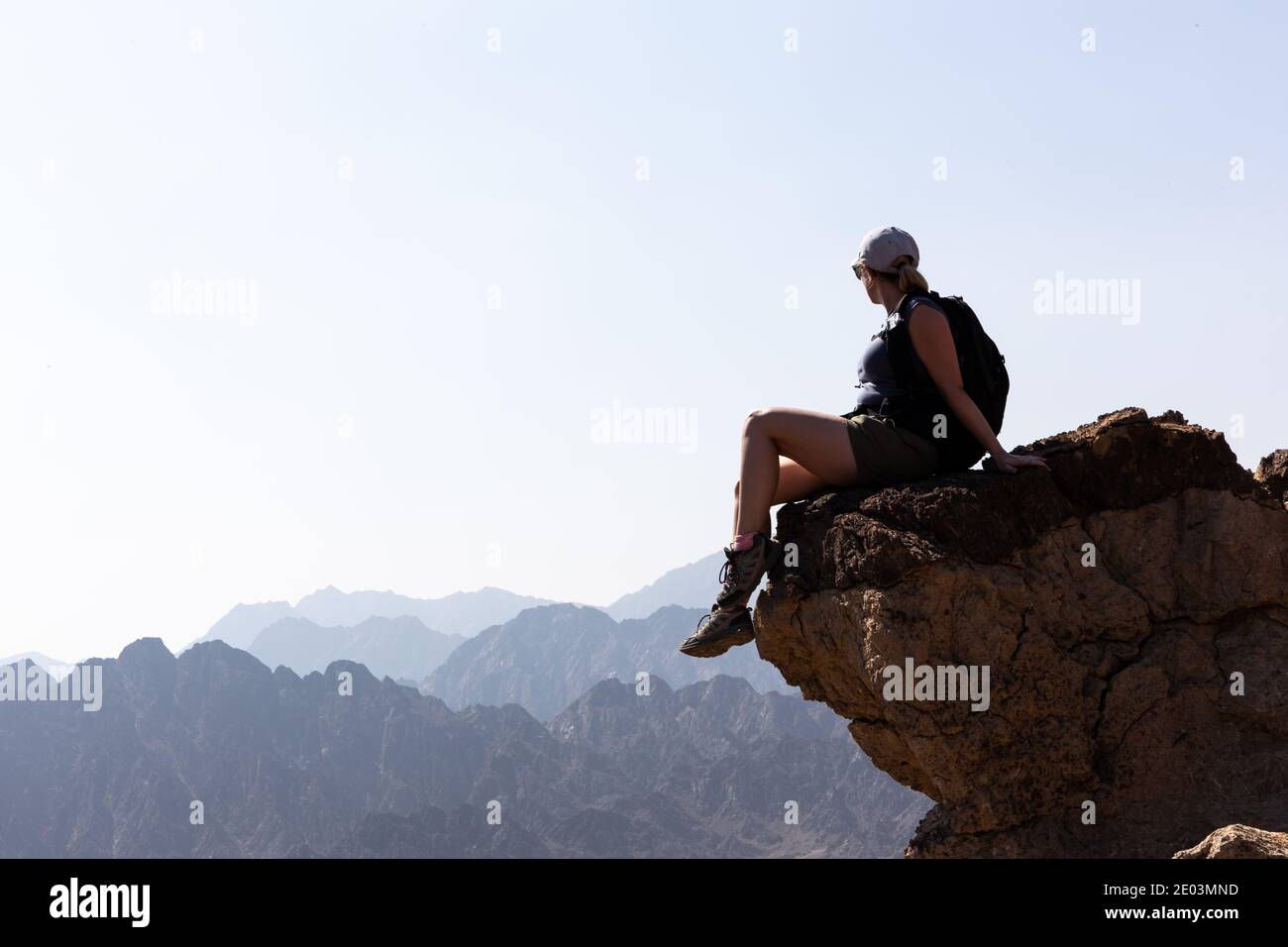 Junge weibliche Tourist Mädchen sitzt am Rande des Felsens mit Abgrund unten, Blick auf die Bergkämme im Horizont, Hatta, Hajar Mountains Stockfoto