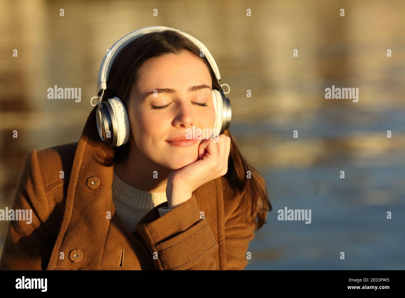 Fröhliche Frau, die Musik hört und Kopfhörer trägt und sich bei Sonnenuntergang entspannt Im Winter am Strand Stockfoto