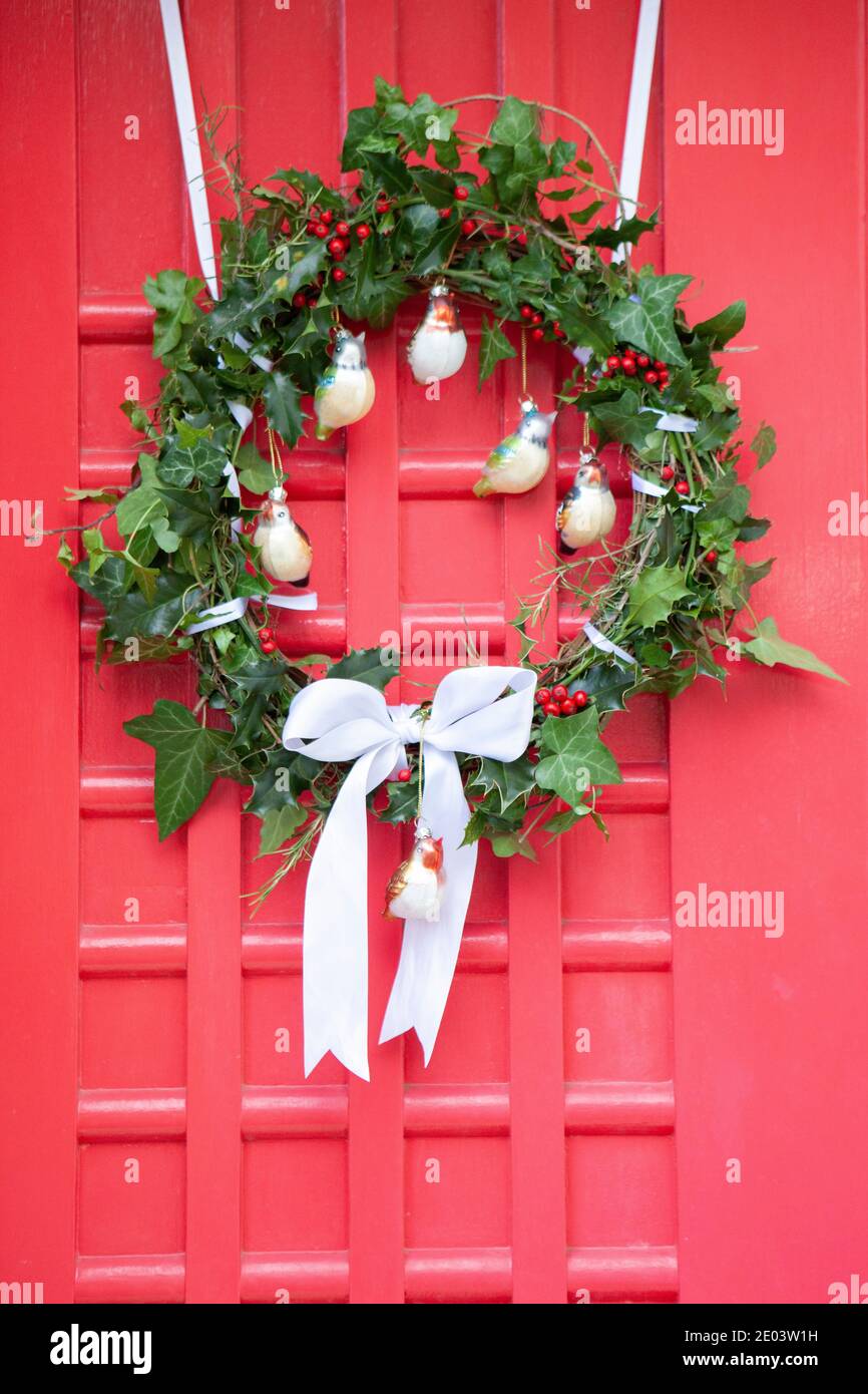 Hausgemachter Weihnachtskranz an einer roten Tür. Der Kranz besteht aus Stechpalme, Efeu und Rosmarinblättern, weißem Band und vogelförmigen Kugeln. Anna Watson/Alamy Stockfoto
