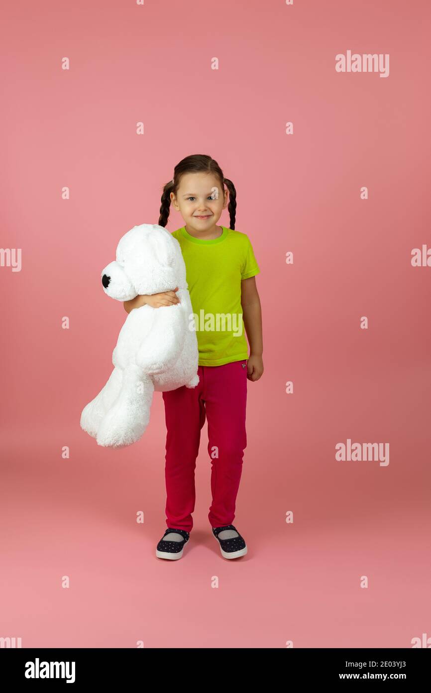 Ein durchgehendes zufriedenes, glückliches pigtailed Mädchen in einem hellgrünen T-Shirt und roter Hose hält einen weißen Teddybär isoliert auf einem rosa Hintergrund Stockfoto