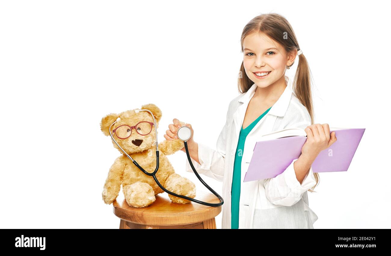 Lächelndes Mädchen trägt in einem medizinischen Mantel mit einem Buch In ihrer Hand spielen Arzt und hören auf ihre Patientin Plüschbär mit Phonendoskop Stockfoto