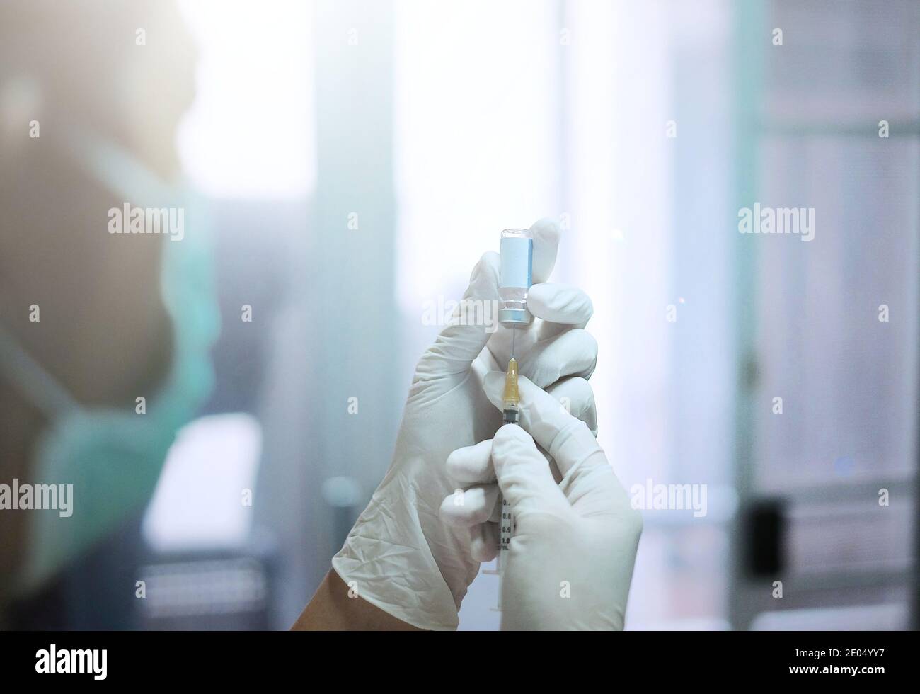 Eine asiatische Ärztin mit weißen Gummihandschuhen und einer OP-Maske in einer Klinik, die eine Nadel und eine Spritze auf eine klare Glasflasche mit Impfstoff einführt Stockfoto