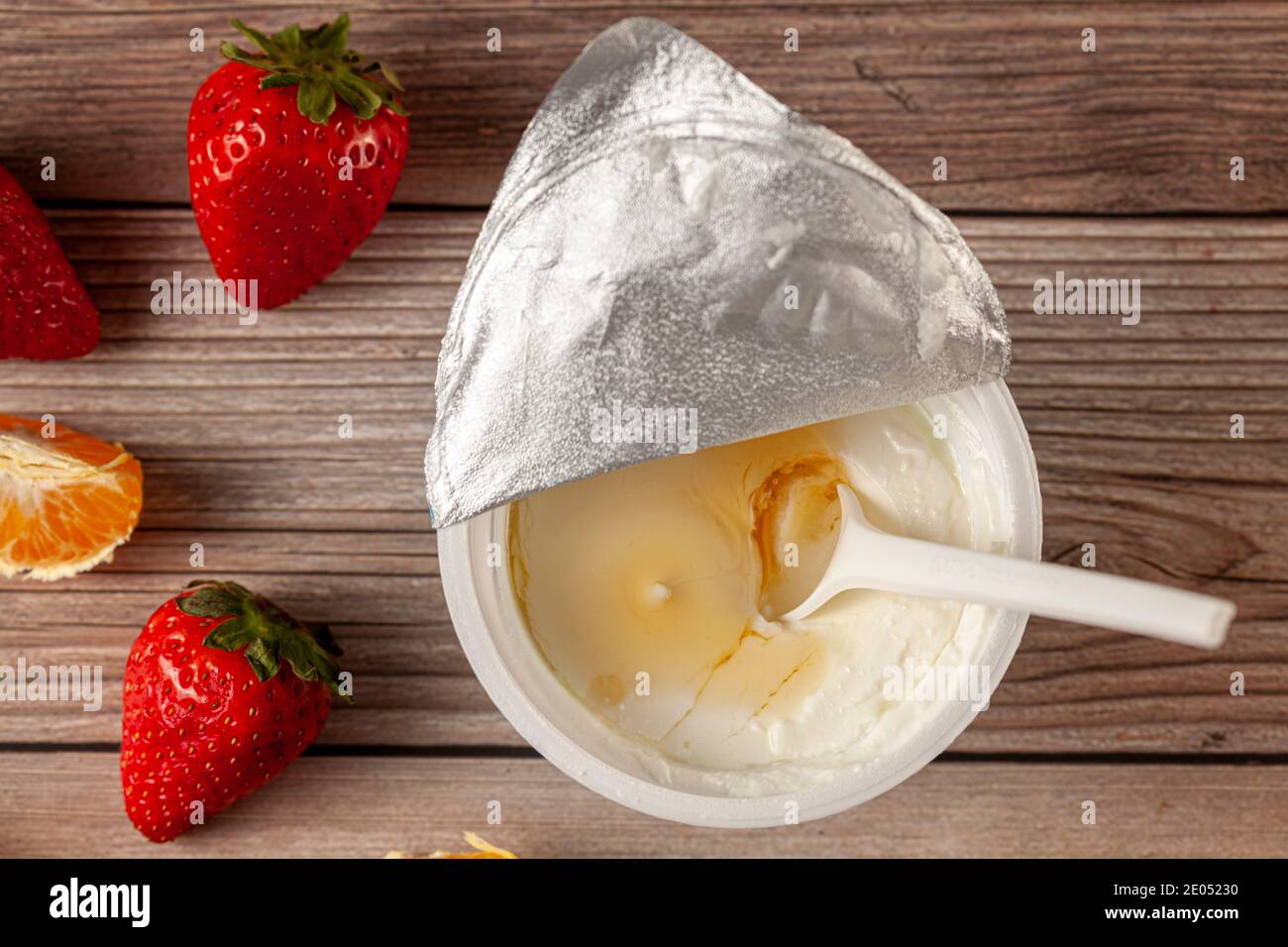 Eine weiße Plastikschale aus reinem griechischem Joghurt mit einem Abgeschälte Aluminiumfolie Dichtung und ein Einweg-Plastiklöffel Eingetaucht serviert mit Mandarinenscheiben A Stockfoto