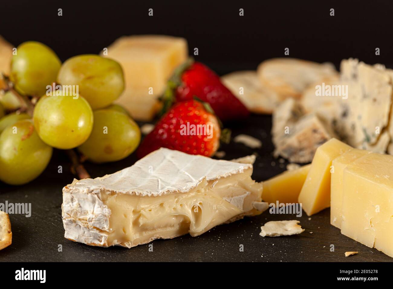 Eine Auswahl an französischen, italienischen und Schweizer Käsesorten in Höhlenalter auf schwarzem Käseplatte, serviert mit Crackern und Früchten (Trauben und Strohhalmchen) Stockfoto
