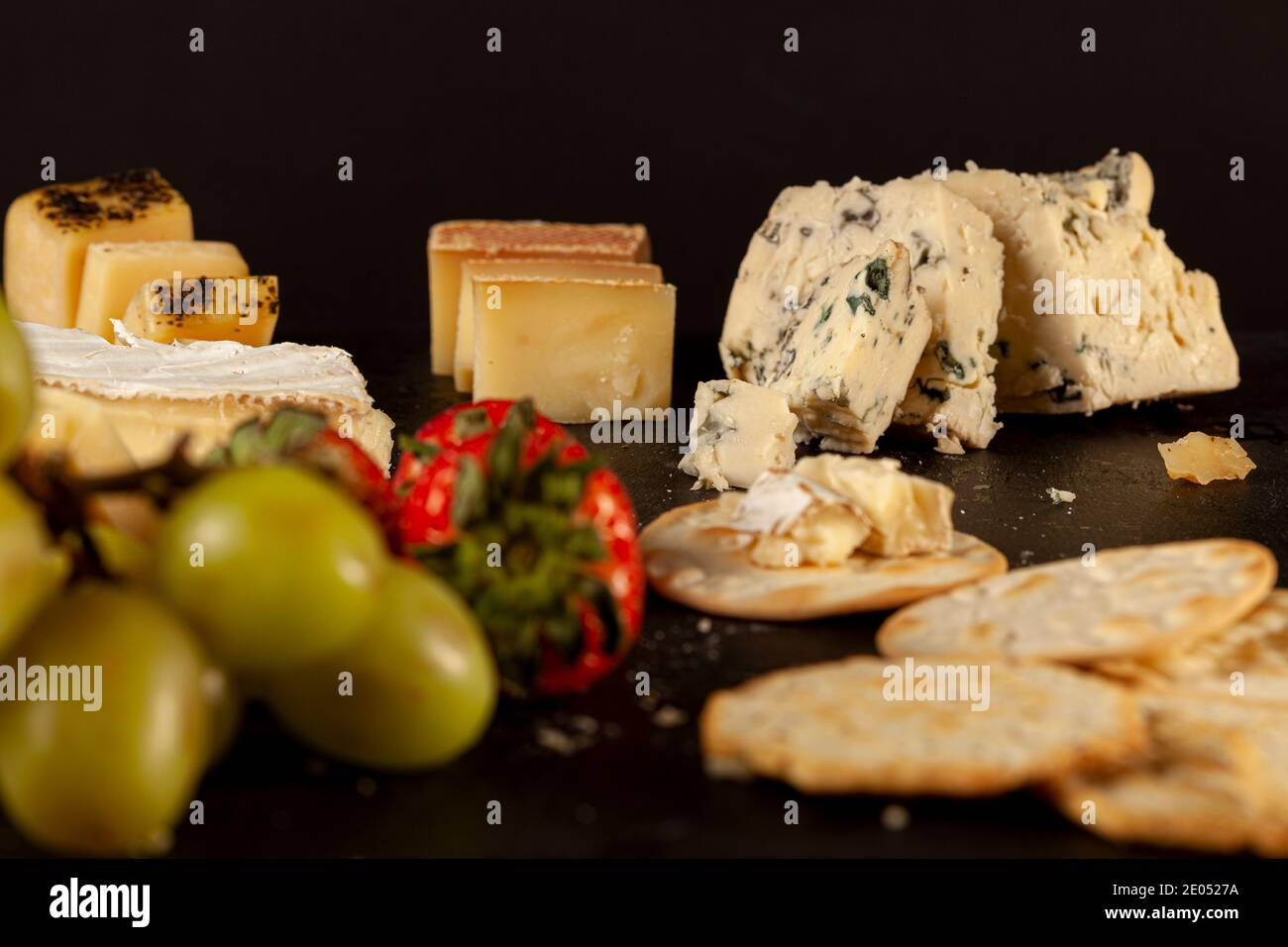 Eine Auswahl an französischen, italienischen und Schweizer Käsesorten in Höhlenalter auf schwarzem Käseplatte, serviert mit Crackern und Früchten (Trauben und Strohhalmchen) Stockfoto