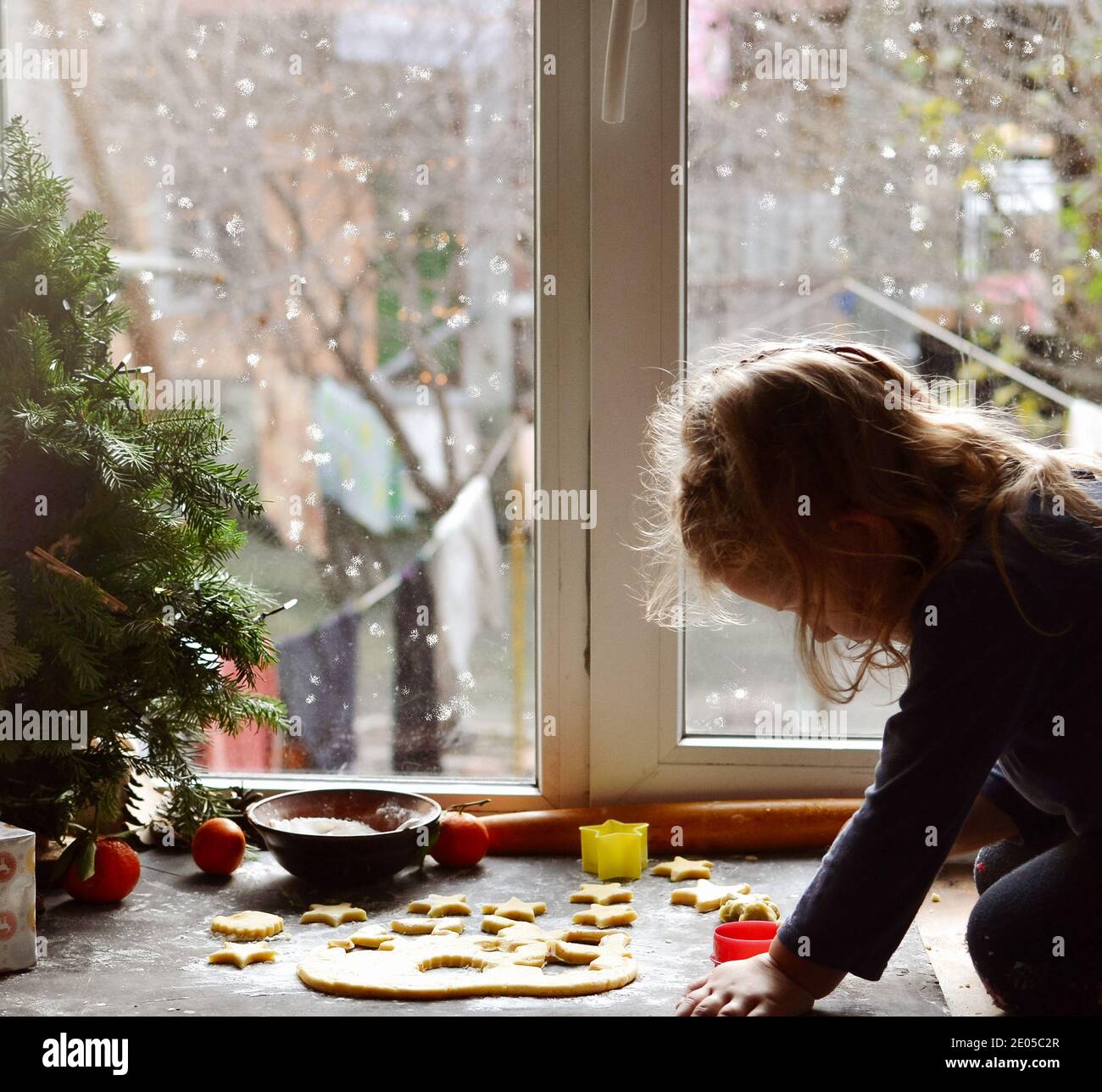 Ein nettes kleines Mädchen 3-4 Jahre alt sitzt auf der Fensterbank und bereitet Weihnachtsplätzchen. Familienurlaub zu Hause während der Ferien. Neujahr und CH Stockfoto