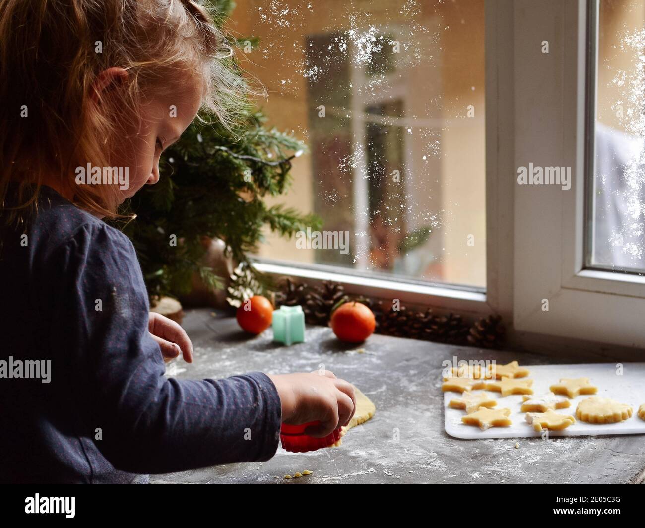 Ein nettes kleines Mädchen 3-4 Jahre alt sitzt auf der Fensterbank und bereitet Weihnachtsplätzchen. Familienurlaub zu Hause während der Ferien. Neujahr und CH Stockfoto