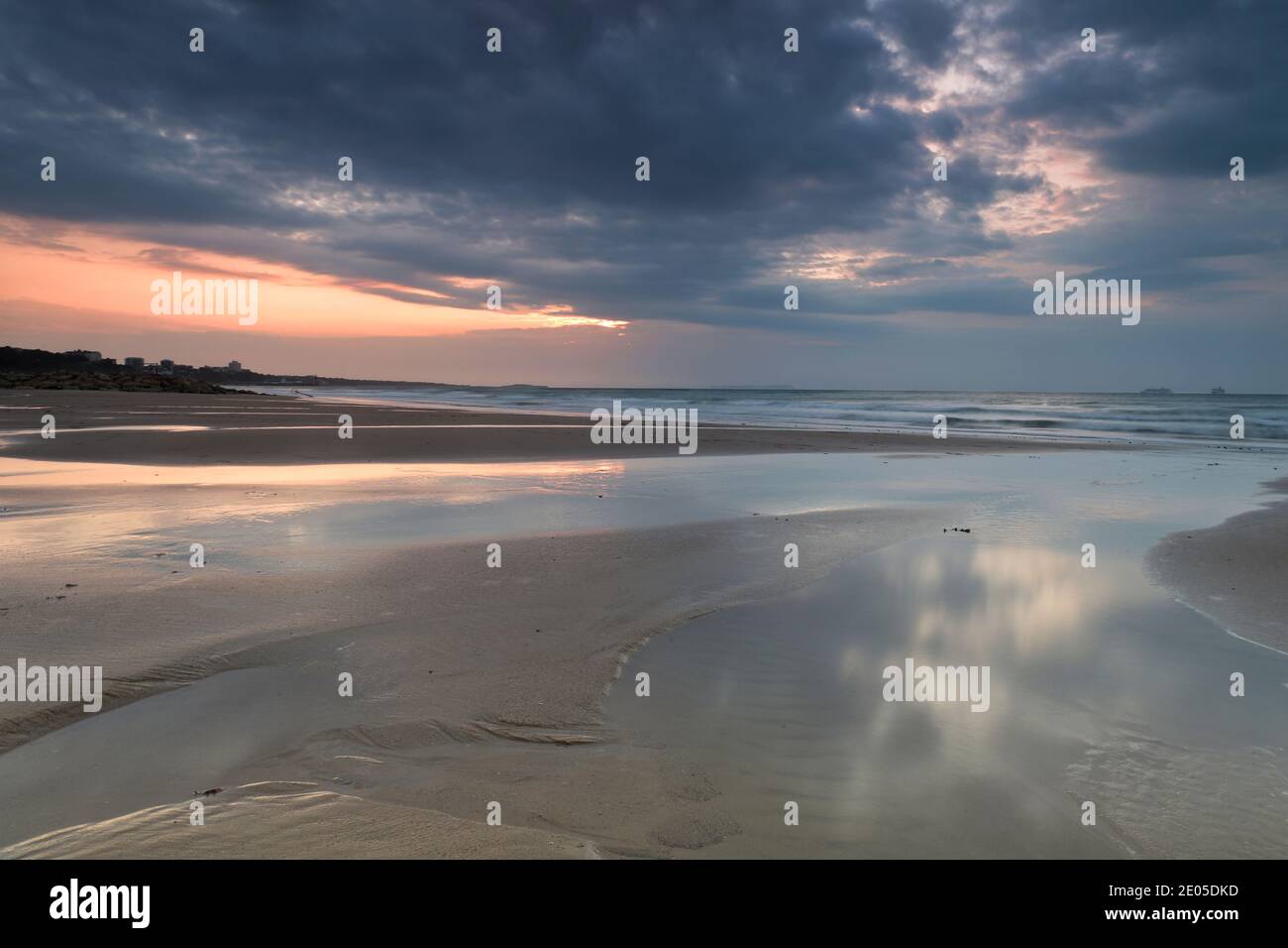 Gezeitenbecken, die vom zurückgehenden Meer in den Sand gehauen wurden, spiegeln den grauen, mit Regenwolken gefüllten Himmel wider, während die aufgehende Sonne über den Horizont kämpft. Stockfoto