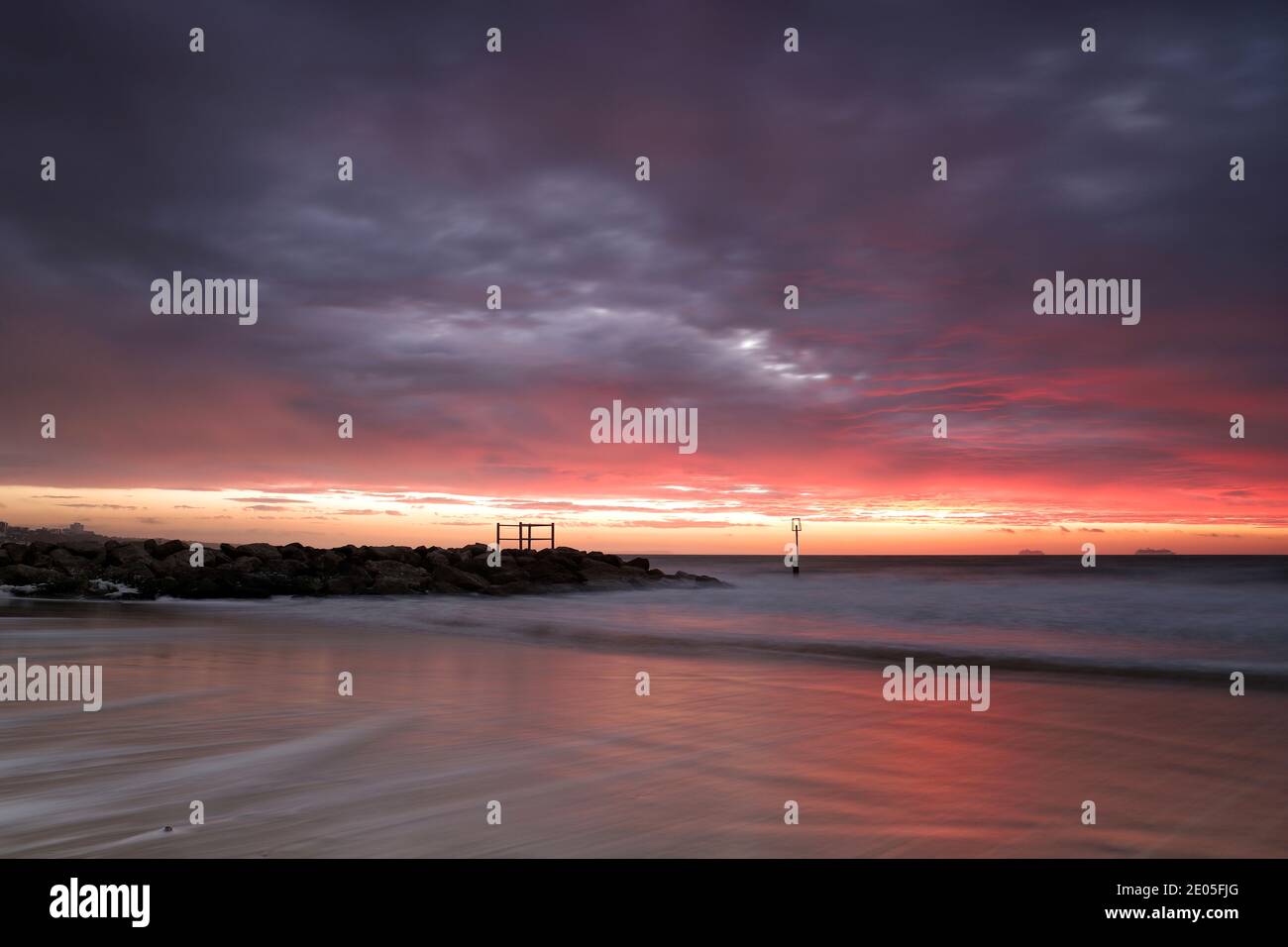 Ein Himmel voller dichter, dunkler Regenwolken ist durch das Licht der aufgehenden Sonne mit Rot durchzogen. Unten Rauschen Wellen über den Sand von Bournemouth Beach. Stockfoto