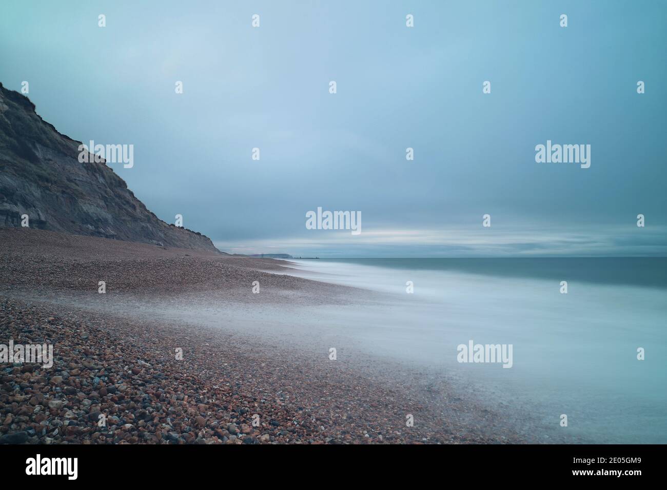 Kieselsteine blicken durch das nebelartige Wasser eines tobenden Meeres, das durch eine lange Belichtungszeit ruhig wurde. Der Winterhimmel ist trüb und voller grauer Wolken. Stockfoto