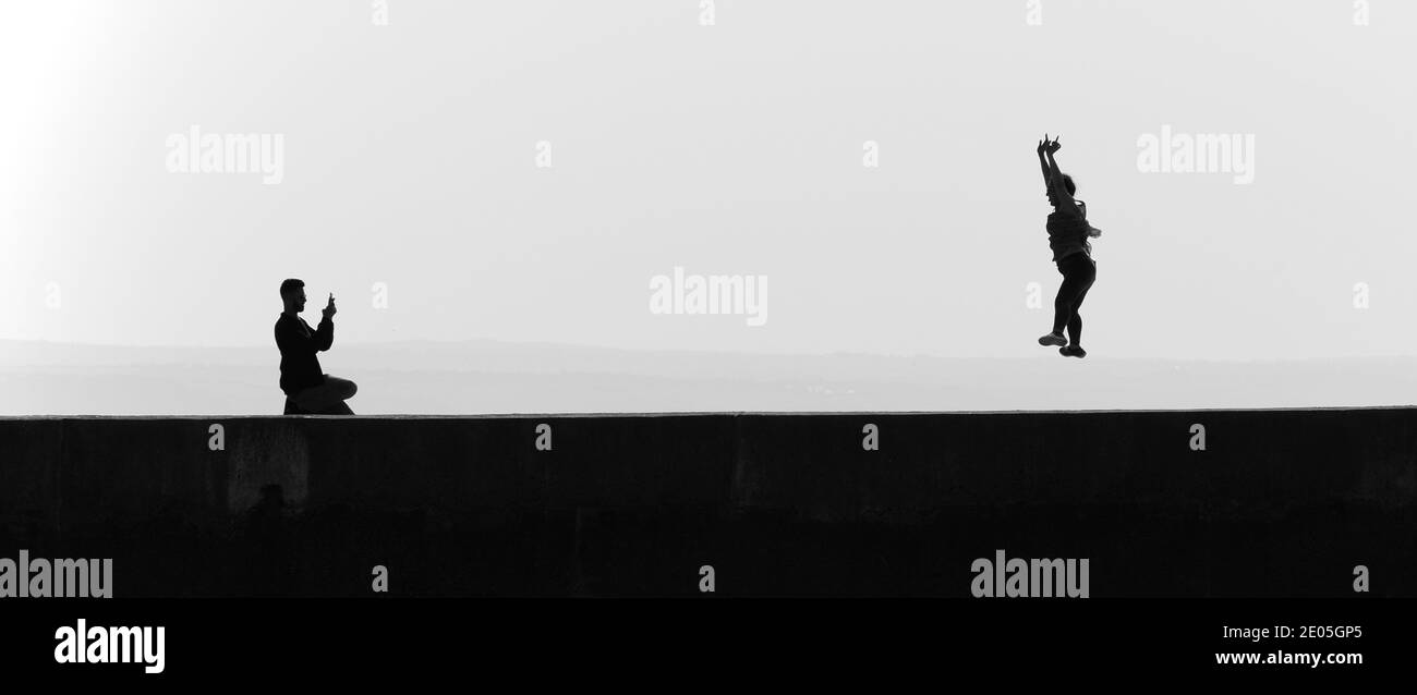 Silhouette eines männlichen Fotografen, der eine Frau fotografiert, die einen Sternsprung macht Mit der Zunge nach außen (S/W) Stockfoto
