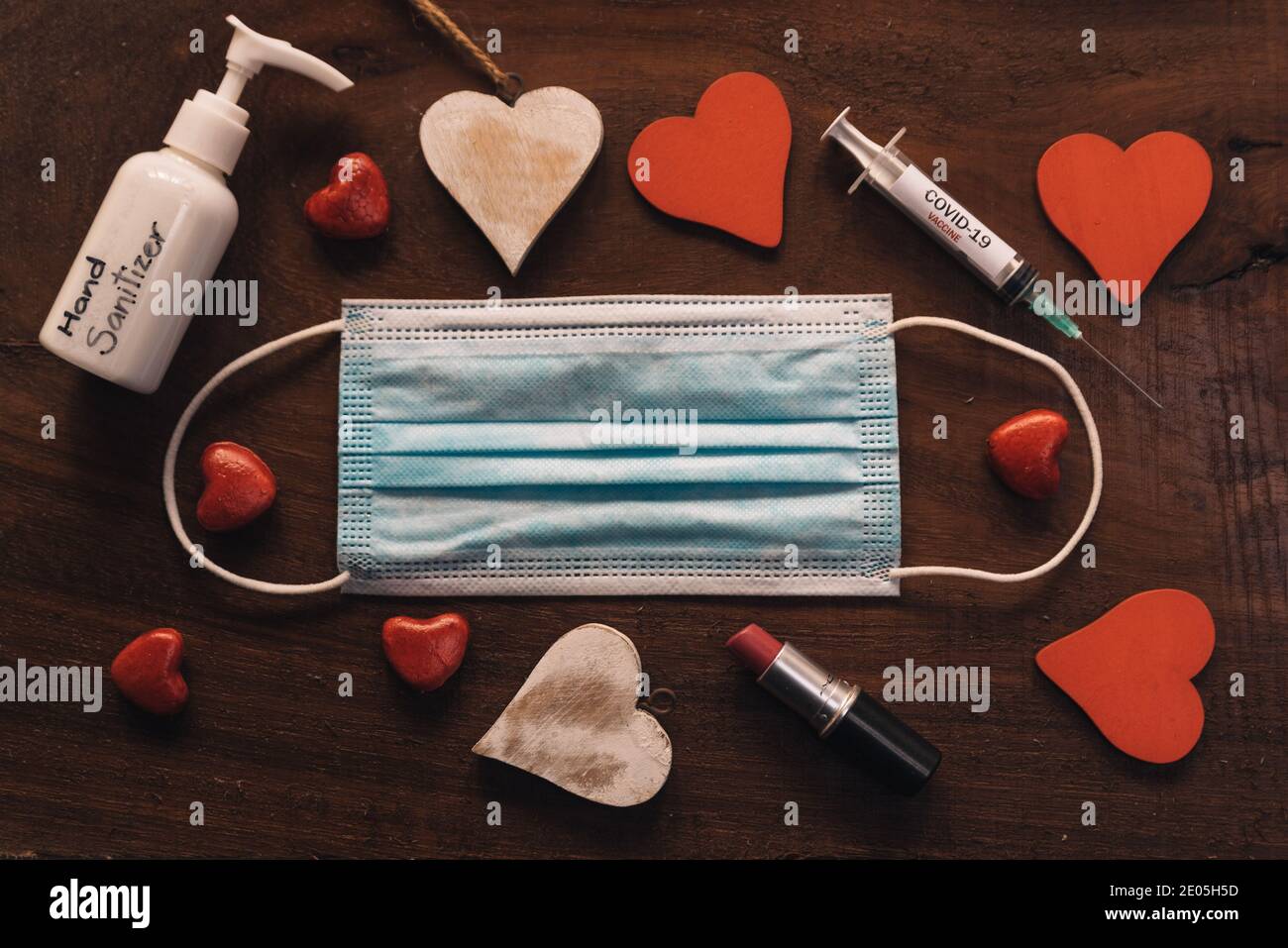 Covid valentinstag Konzept. Gesichtsmaske mit roten und weißen Herzen auf Holz Hintergrund. Draufsicht der Hand Desinfektionsmittel und Lippenstift und Spritze für die n Stockfoto