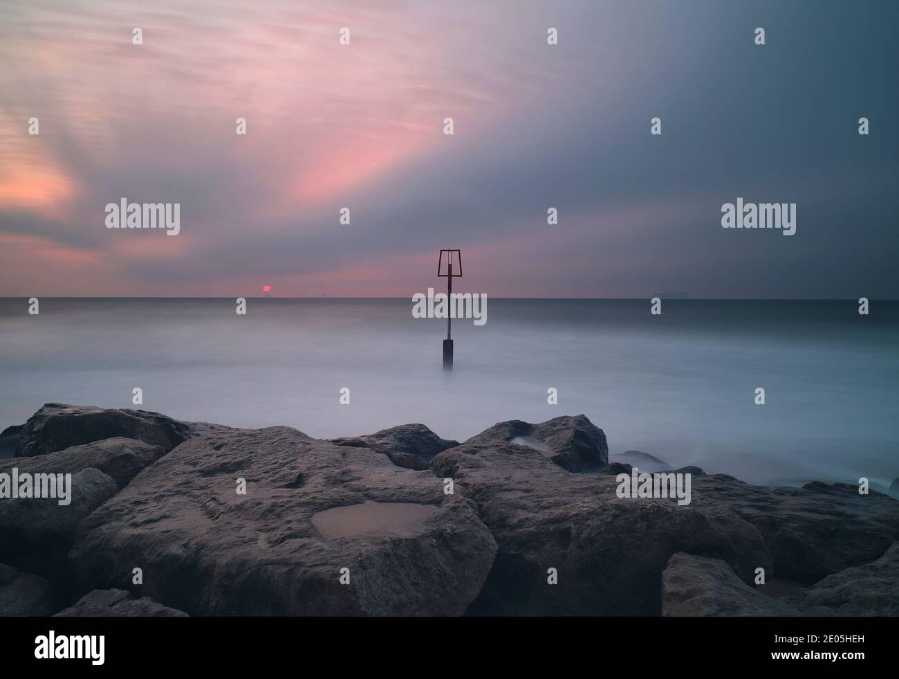 Die Sonne beginnt in einen Himmel voller trübe Wolken aufzusteigen. Ein einziger Groyne-Pfosten erhebt sich in der Mitte des Bildes aus einem durch eine lange Belichtung beruhigten Meer. Stockfoto