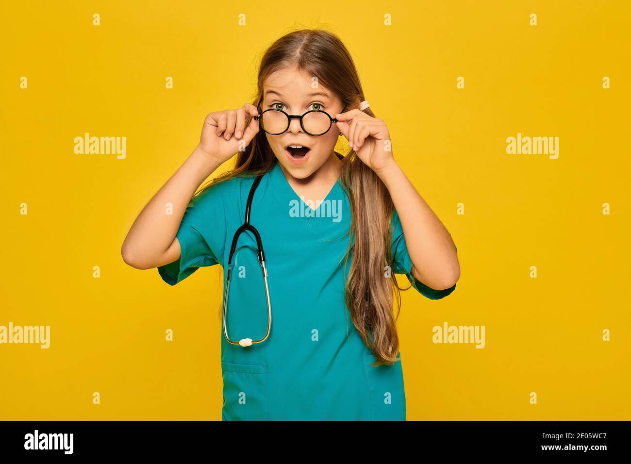 Kind trägt in einer blauen OP-Uniform und einem Stethoskop spielt den medizinischen Beruf, und zeigt auf ihrem Gesicht Gefühl der Überraschung, das Ausziehen von Glas Stockfoto