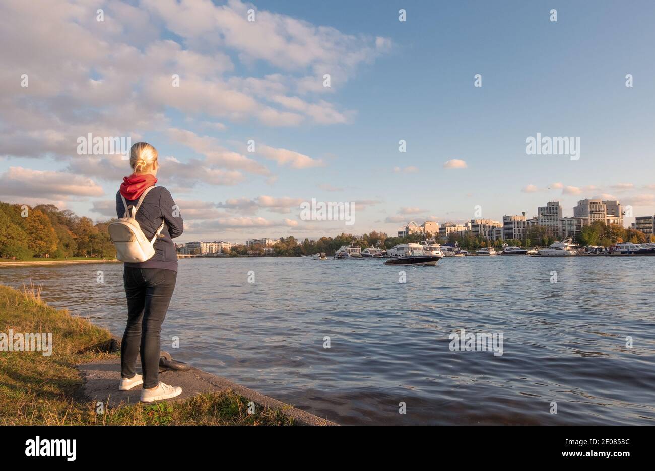 Eine junge Frau in lässiger bequemer Kleidung und mit Rucksack steht am Flussufer und bewundert die Aussicht. Alleinreise, lokales Tourismuskonzept. Stockfoto
