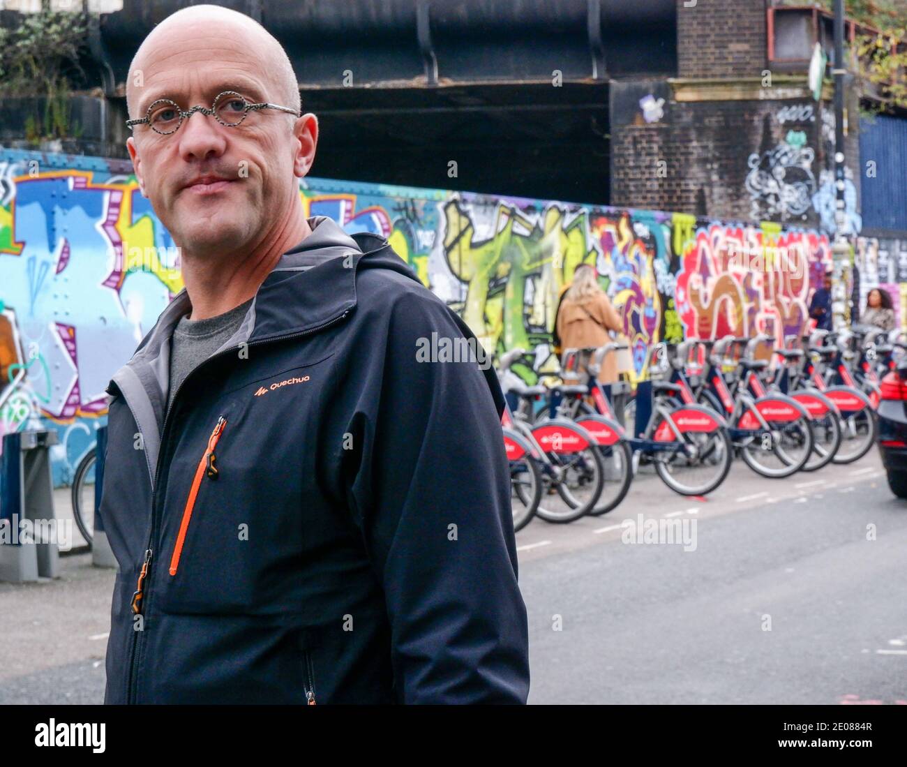 Ein Mann mit kleinen runden Gläsern läuft vor Santander Bikes Docking Station & Graffiti Wand auf Brick Lane, Shoreditch, East London. Stockfoto